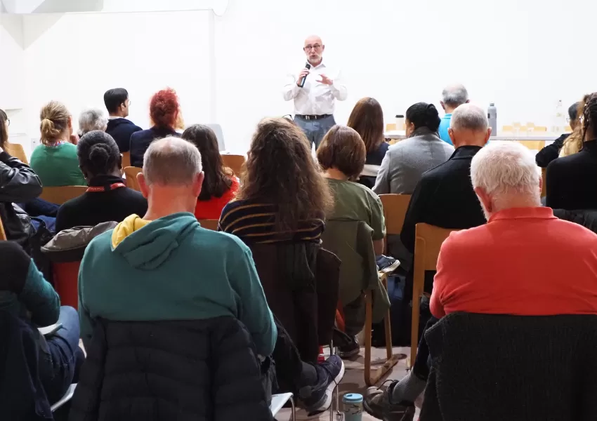 The MC stood at the far end of the room, looking at the camera over the heads of his seated audience 