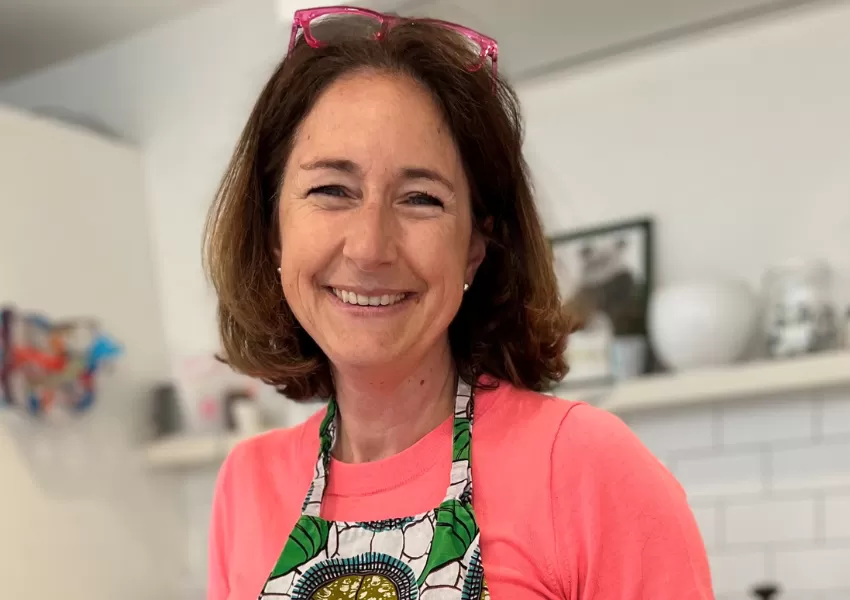 Kat smiling to camera, wearing a pink jumper and a green floral patterned kitchen apron
