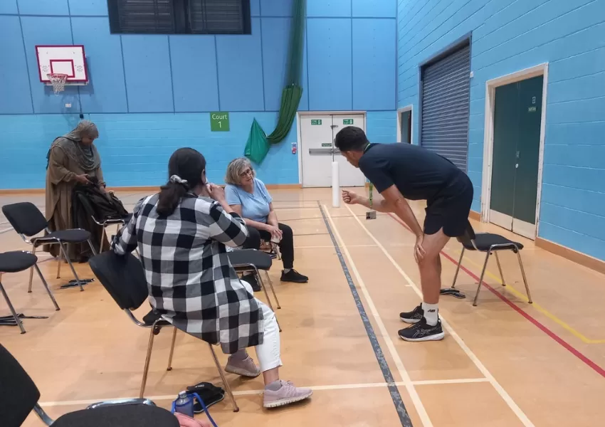 Two women sat down on chairs in the gym, with cups of tea, talking to a male fitness instructor