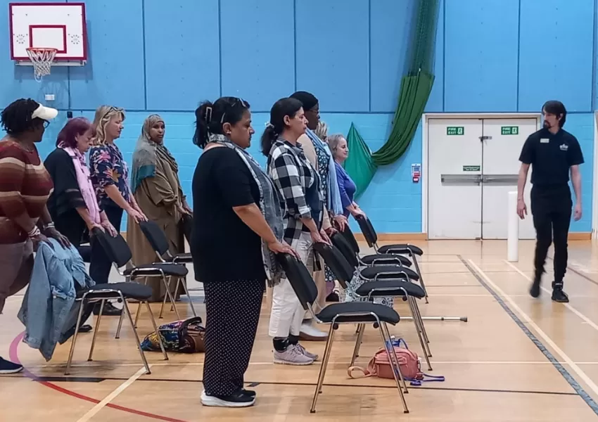 Rows of people, stood behind chairs, looking to the front of the gym at their instructor