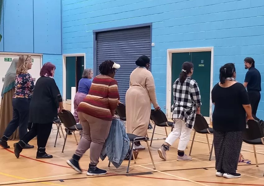 Rows of people stood behind chairs in a gym, lifting legs behind them, with instructor at the front