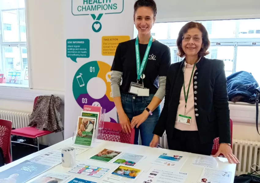 Two women smiling at a table with health leaflets. Sign reads Barnet Health Champions. 