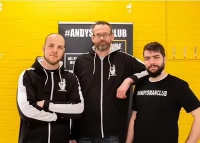 three men wearing black and white 'Andy's Man Club' sweaters, stood together against a yellow brick wall