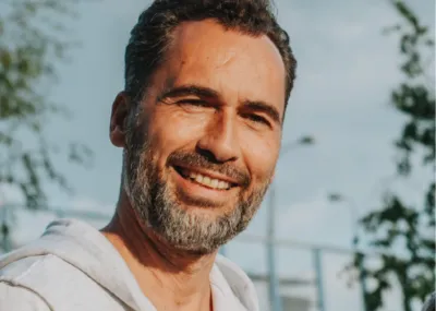 Middle aged man with dark hair and silver beard, smiling with sunshining on his face. Outside with a blue sky behind him, wearing a grey hoody.